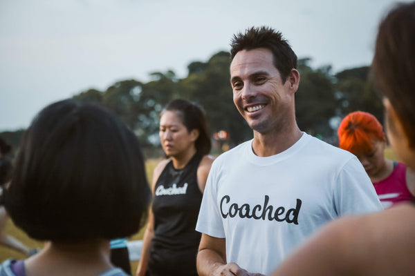 A coach wearing a white 'Coached' t-shirt interacting with his runners outdoors.